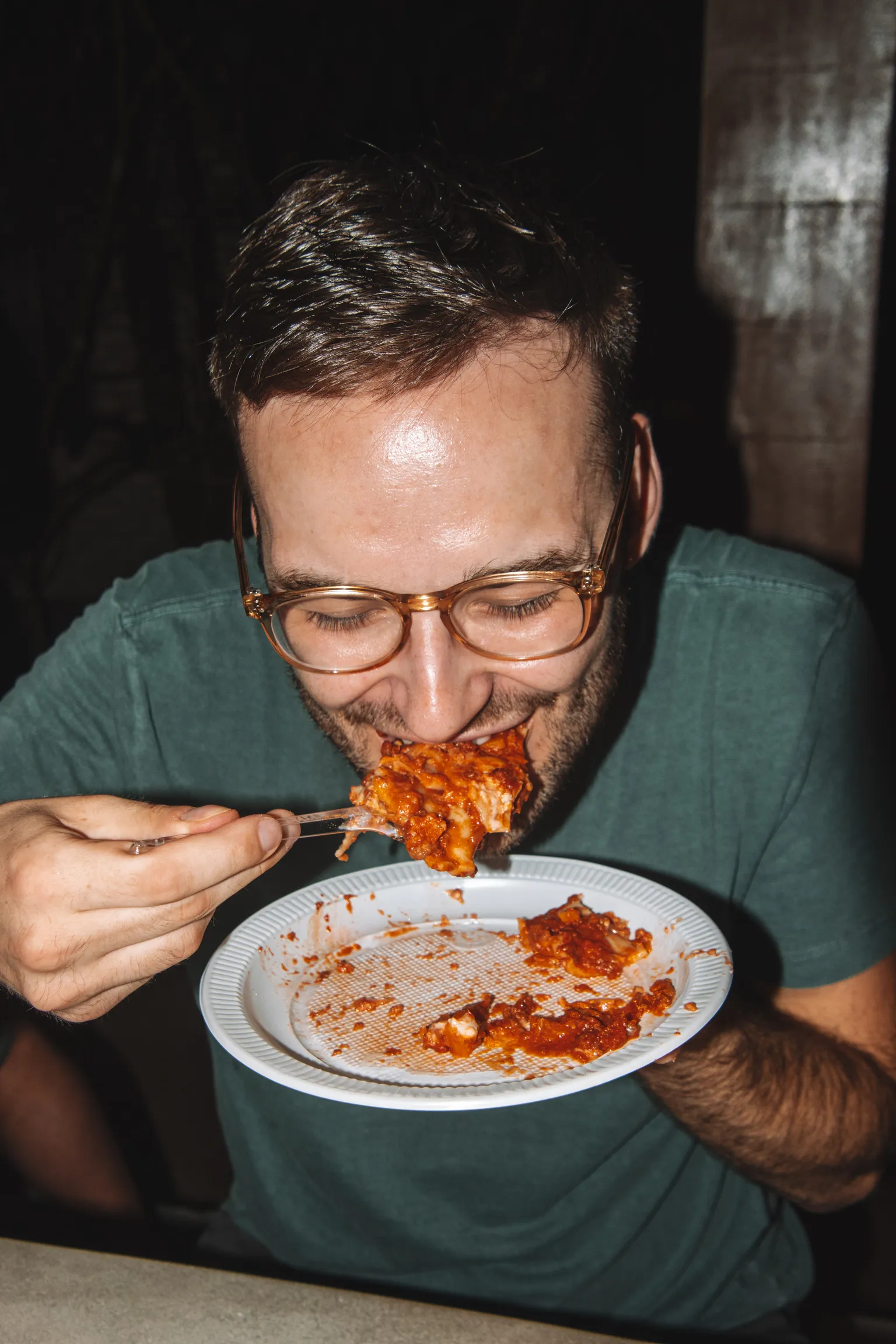 Man wearing glasses and a green shirt eats a forkful of lasagna from a white paper plate.